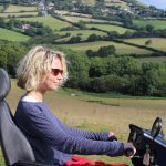 woman wearing sunglasses on an all terrain mobility scooter, also known as tramper, with green hills and countryside behind her, she looks happy, it is a bright sunny day.