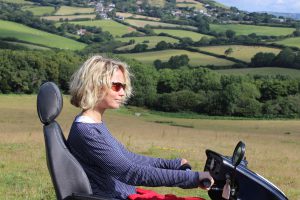 woman wearing sunglasses on an all terrain mobility scooter, also known as tramper, with green hills and countryside behind her, she looks happy, it is a bright sunny day.