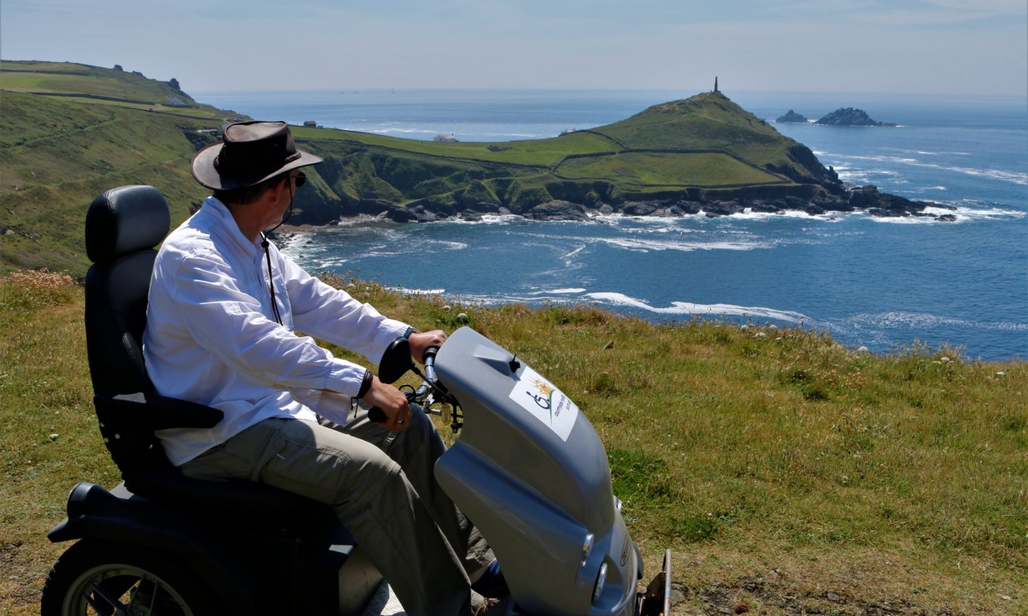 Man wearing a hat on a sunny clear day is seated on a stationary all terrain mobility scotter, looking away from camera and takin in the costal view, there's an obelisk in the distance n the coastal head with waves rolling into the bay below.