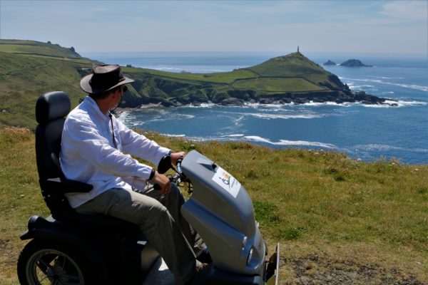 Man wearing a hat on a sunny clear day is seated on a stationary all terrain mobility scotter, looking away from camera and takin in the costal view, there's an obelisk in the distance n the coastal head with waves rolling into the bay below.