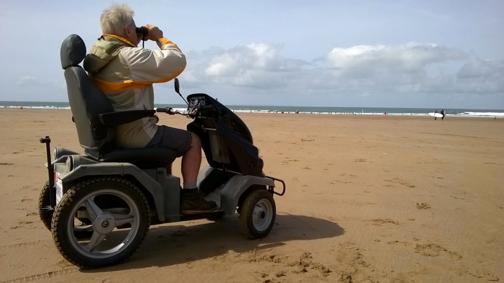 Man looking through binoculars onn the beach seated in an all terrain mobility scooter