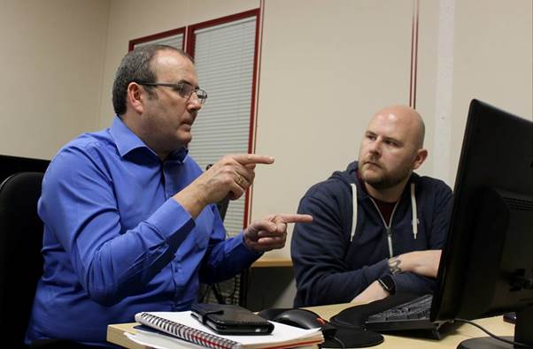 Two men seated at desk facing computer screen signing in Brtish Sign Language