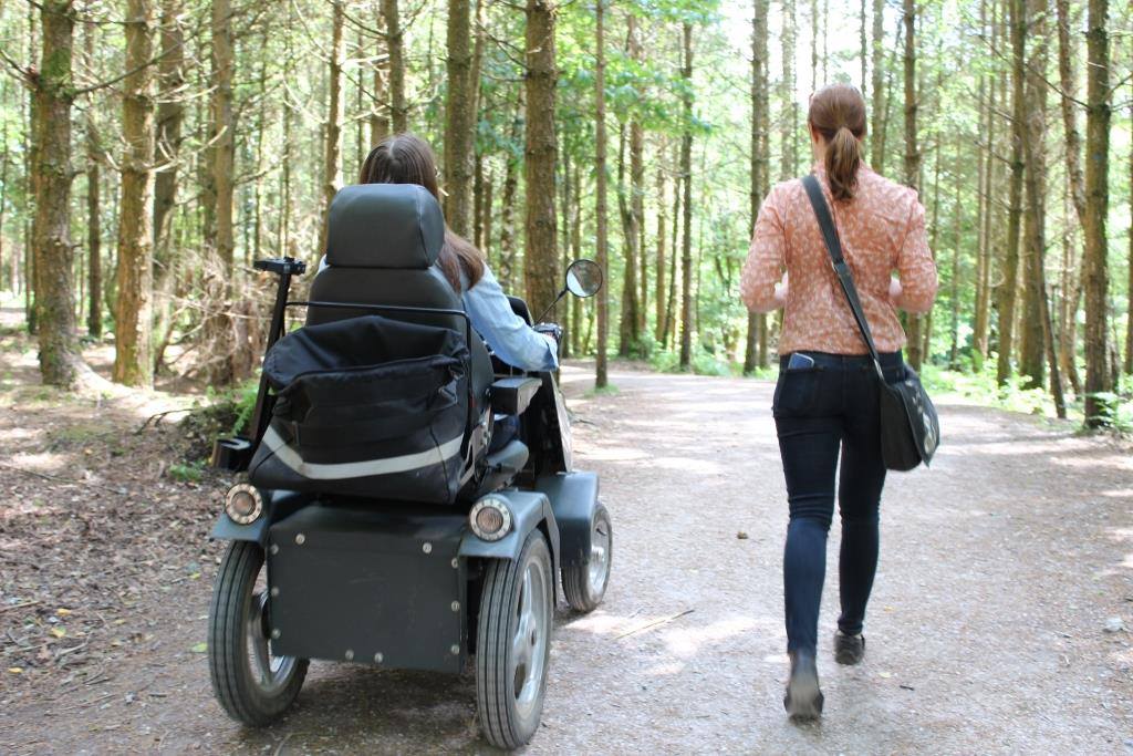 Two people in the outdoors, a green wood on a sunny spring summer day, the person on he left is in an all terrain mobility scooter and the other person is walking alongside.