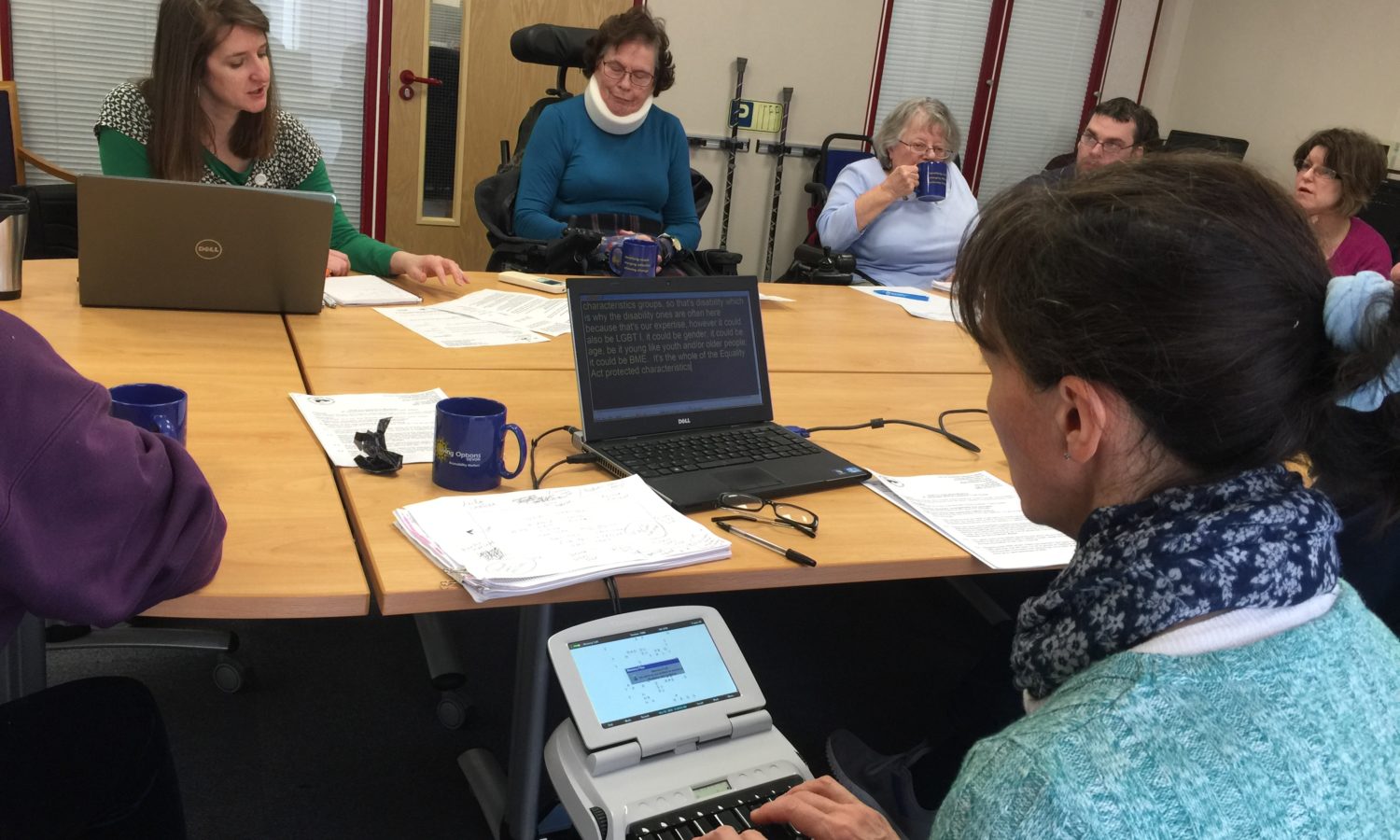 People seated around a desk in the Living Options Devon meeting room, in the forefront is a lday with her back to us typing speech to text, across the desk are three women seated in wheelchairs, drinking tea and taking part in the meeting with three others seated around the desk.