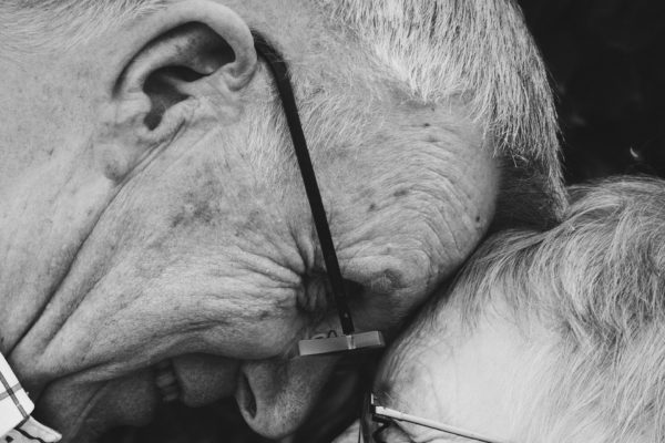 An elderly woman and man wearing glasses, heads together, smiling at each other.