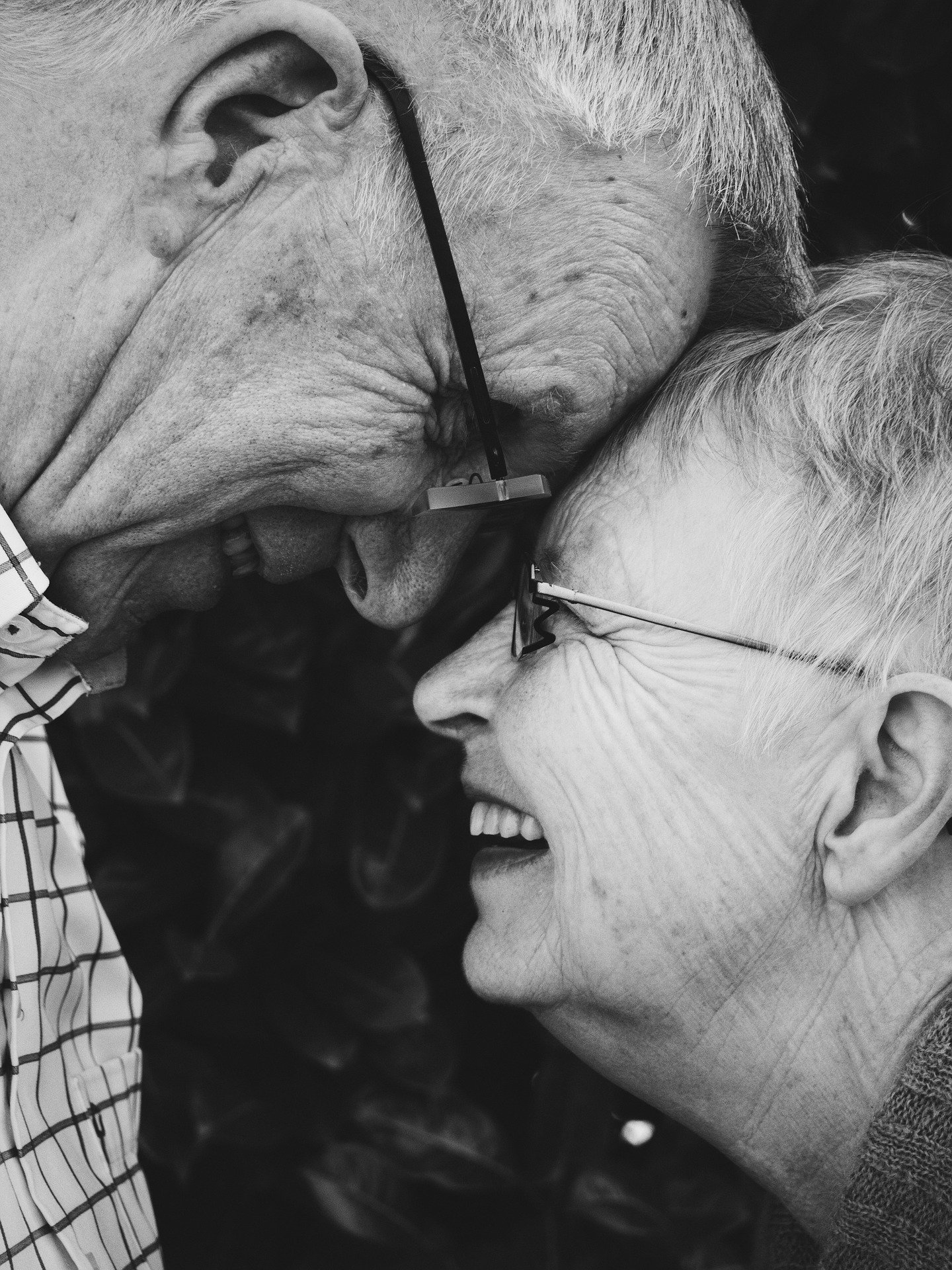 An elderly woman and man wearing glasses, heads together, smiling at each other.