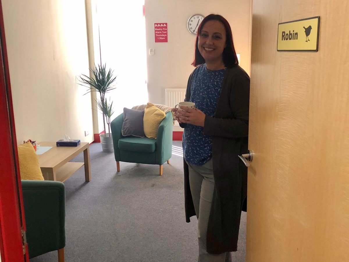 Smiling woman stood next to a door into a bright room with pot plant and soft furniture.
