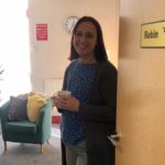 Smiling woman stood next to a door into a bright room with pot plant and soft furniture.