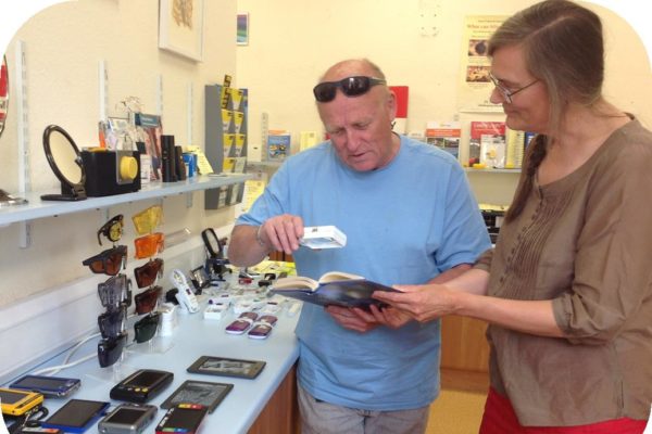 A room with different types of equipment to help you see and hear if your have a sensory loss impairment. A man is stood with a woman and uses a magnifier to read a book.