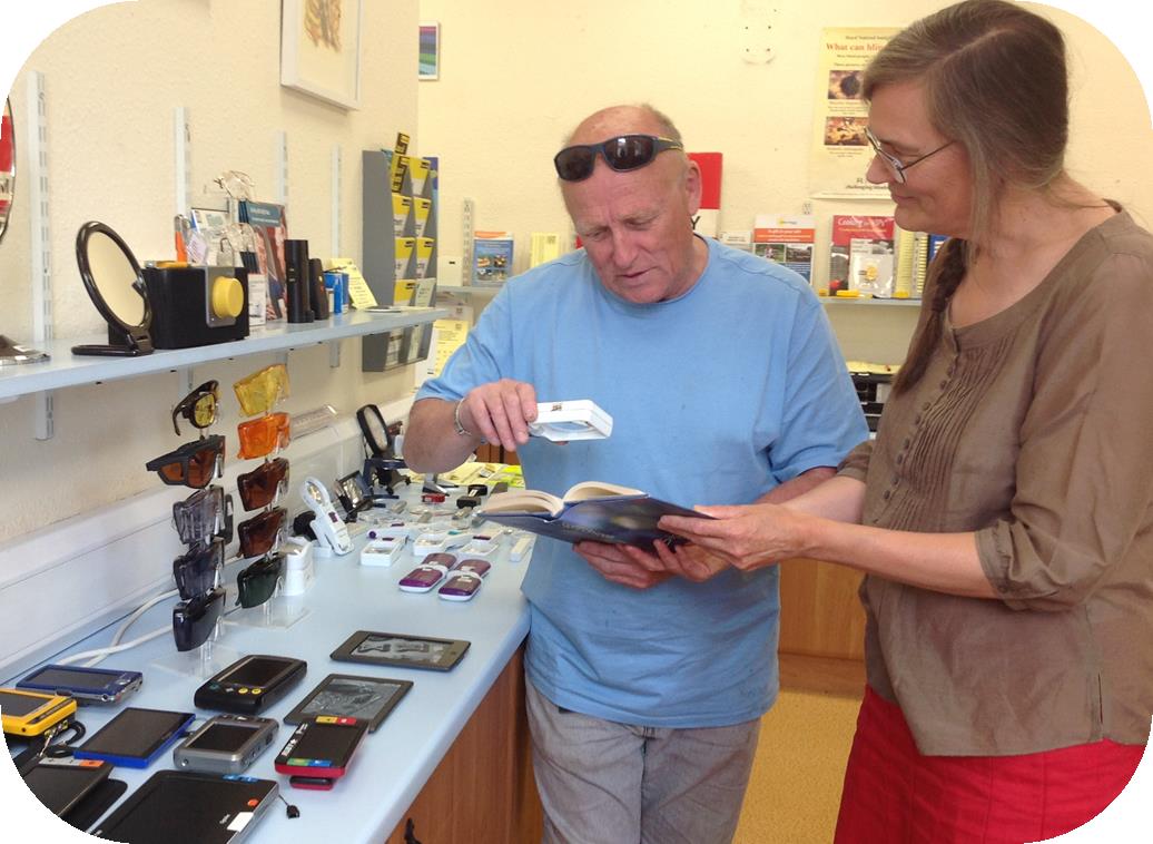 A room with different types of equipment to help you see and hear if your have a sensory loss impairment. A man is stood with a woman and uses a magnifier to read a book.