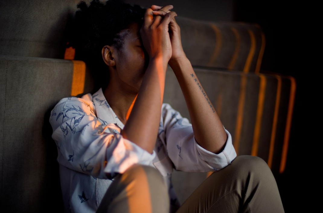 Woman sat in the dark with stripes of orange light coming through the window on her. She has her head in her hands, lonely and sad