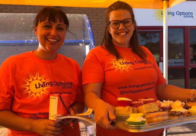 Two members of Living Options Devon with cakes and a donation bucket under the Living Options Devon gazebo at an event