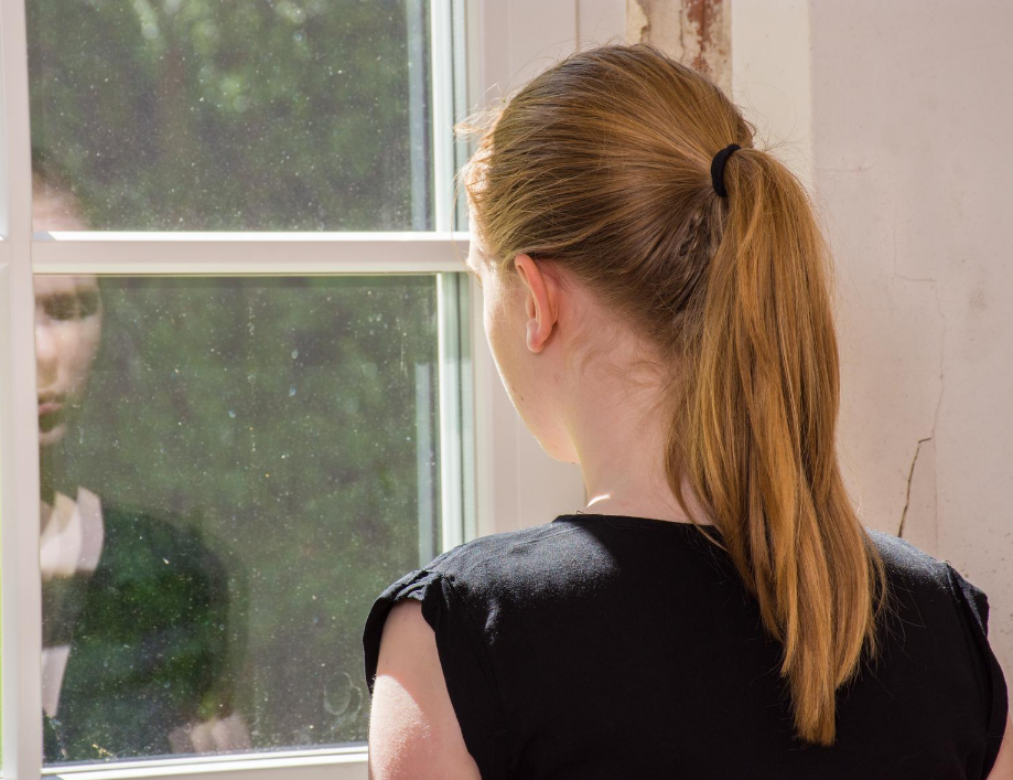 A young lady with blonde hair in a ponytail, wearing bacl top, stares out of a window, it's raining outside and we see her sad face refelcted back.
