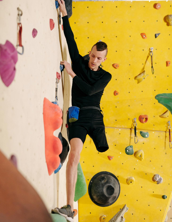 Man wearing black top and black shorts, he is missing his left leg, he is facing camera on an indoor climbing wall which is yellow with colourful holding for your feet and hands.