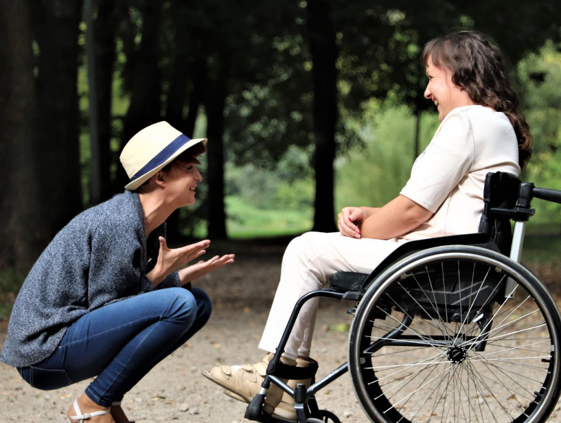 Two women in an outdoor space, one is kneeling down laughing with the other woman who wears white and is in a wheelchair laughing