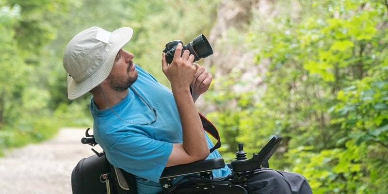 wheelchair user holding camera taking photos outside in a green area. He wears a hat and blue t-shirt