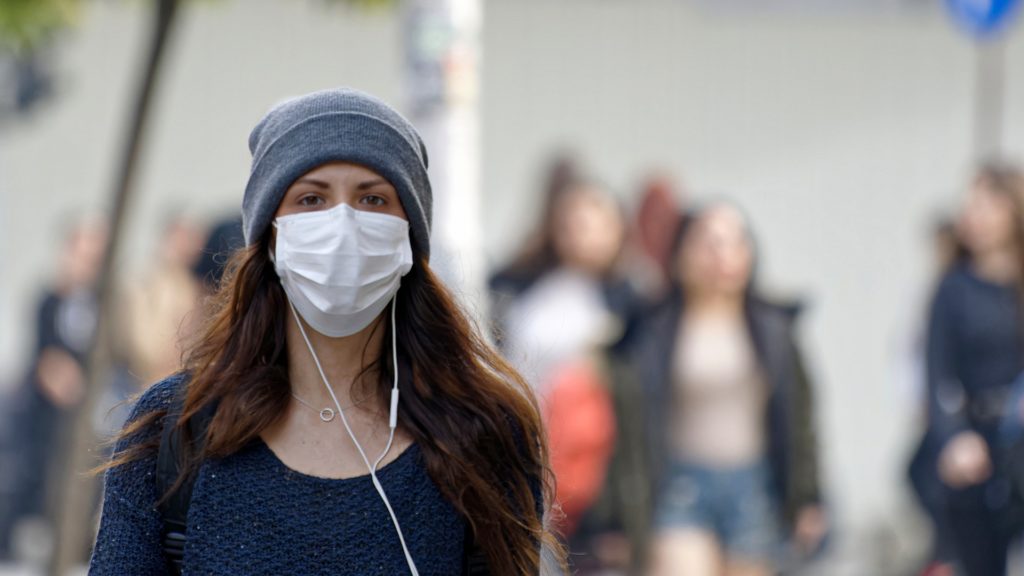 woman in street walking wearing a face mask and listening to music. She has long hair and a wooly hat, people blurred walking behind her.