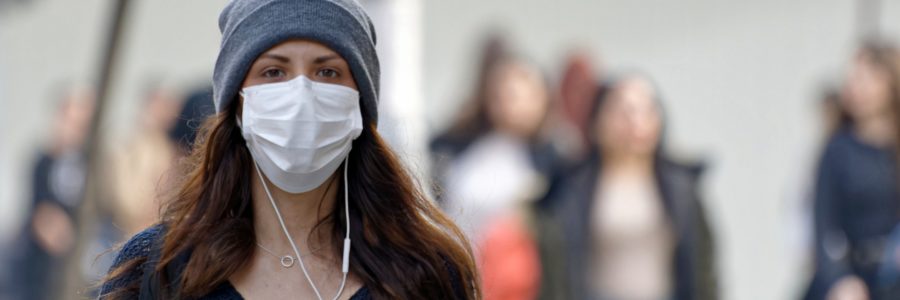 woman in street walking wearing a face mask and listening to music. She has long hair and a wooly hat, people blurred walking behind her.