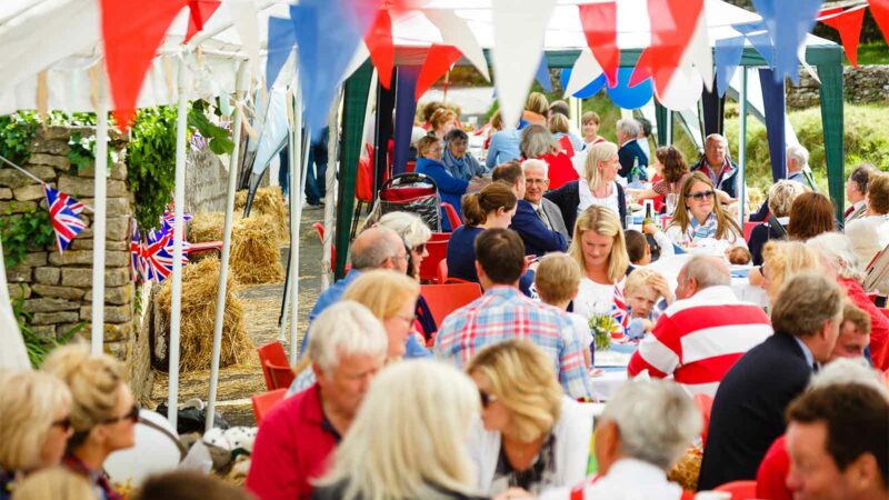 A street party with lots of people at tables celebrating with red, white, and blue flags