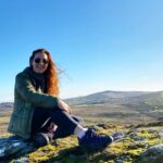 Becki Haddock, sitting on the top of a Dartmoor tor in the sun with blue sky behind