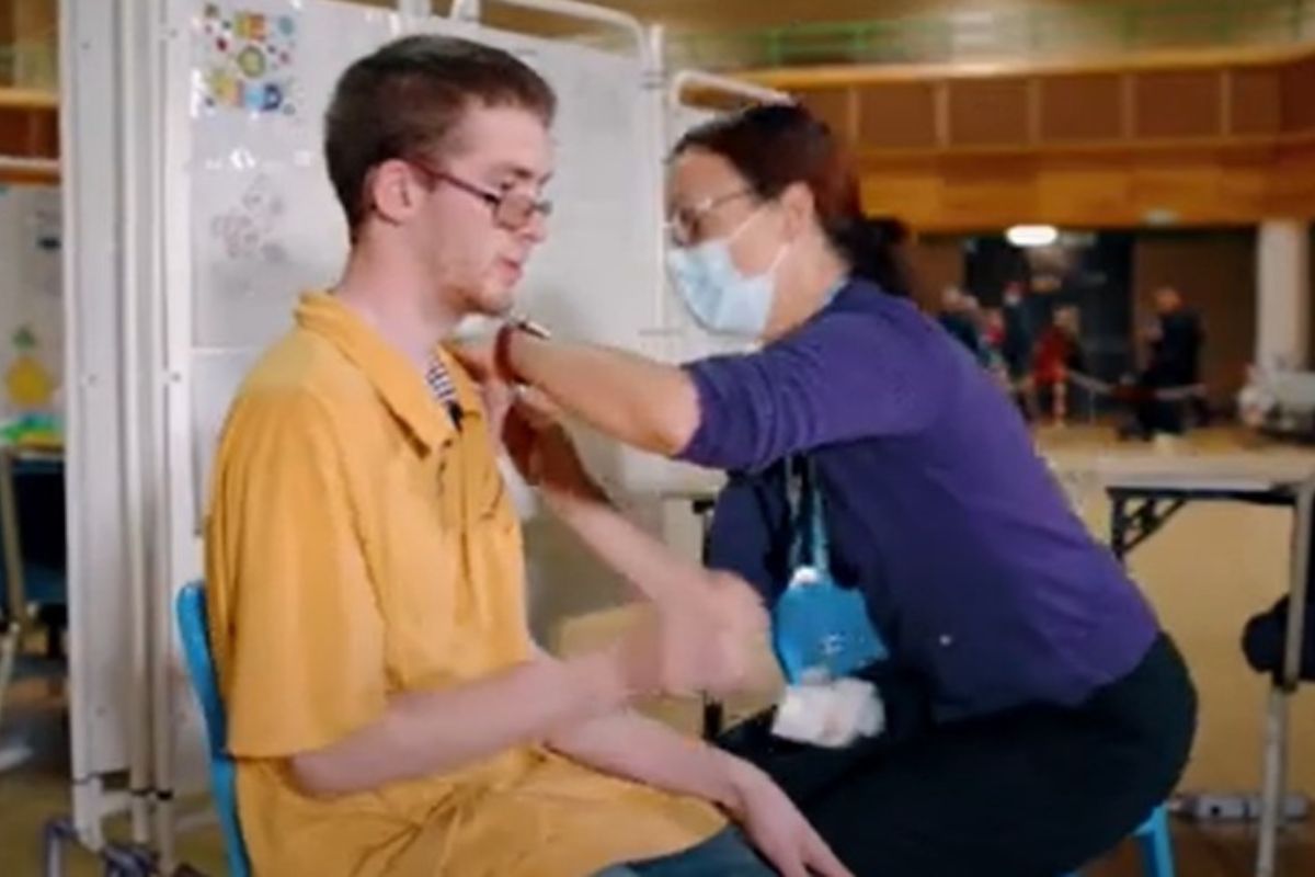 A male sat in a chair receiving an injection by a female wearing a face mask and lanyard
