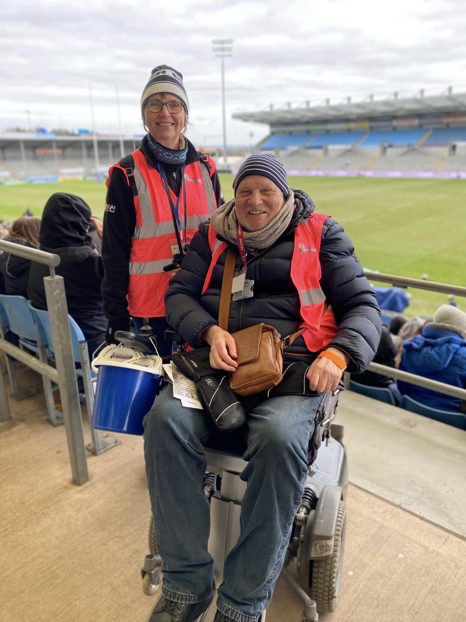 Man in power assisted wheelchair and women standing by a rugby pitch both in high-vis tabards