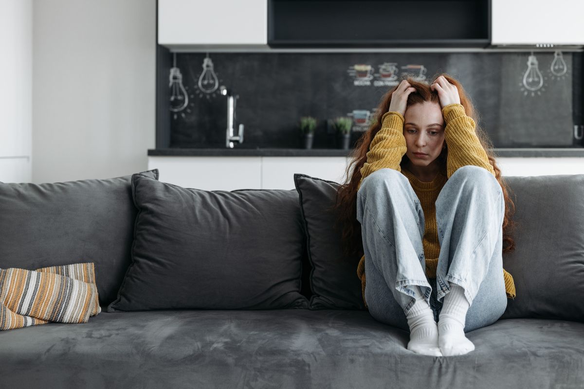 A person holding their head in their hands sitting on a sofa with knees tucked up