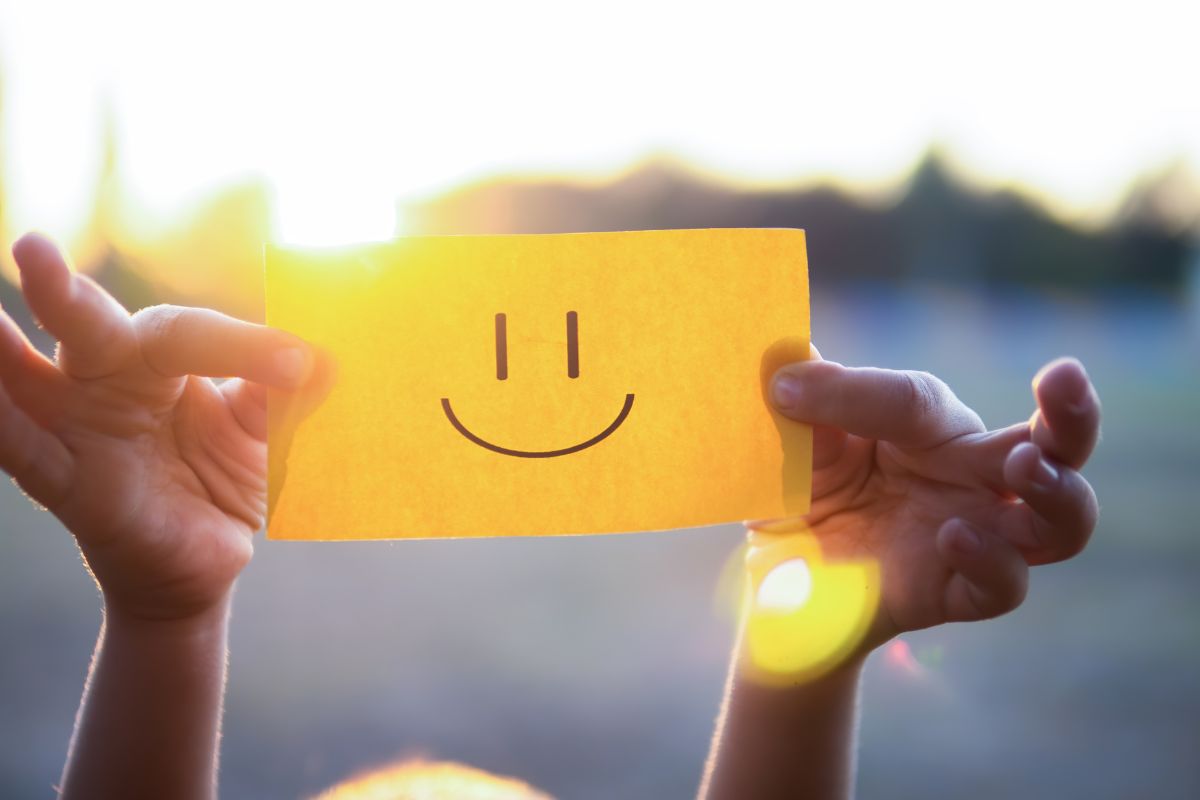 A hand-drawn smiley face on a yellow sticky note being held by two hands against bright sunlight