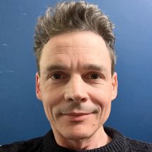 As smiling male looks straight at the camera against a blue background and wearing a black coloured top just visible