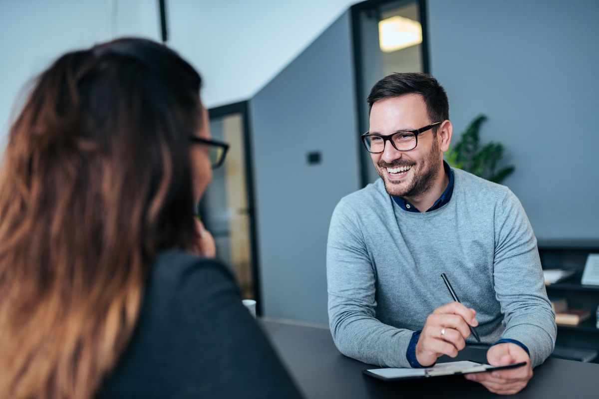 Two people sit in an office environment looking directly at each other. The person in view looks relaxed and smiles.