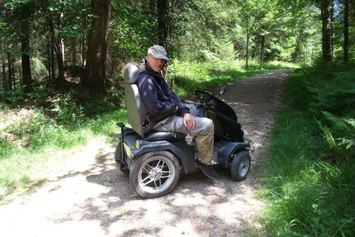 Steve sits in a Countryside Mobility tramper. Sunlight shines in between the tree lined path