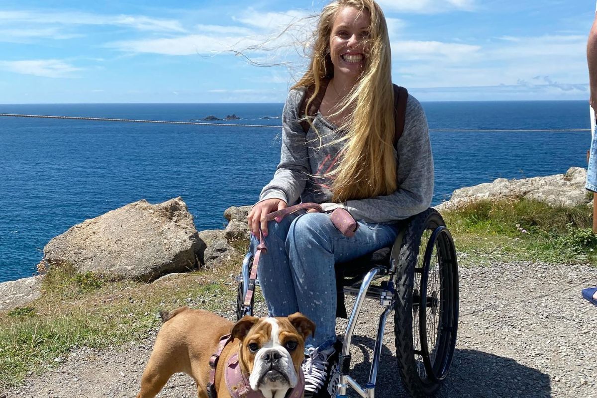 Casey sits with her dog on a lead with the sea and coast in the background on a bright sunny day