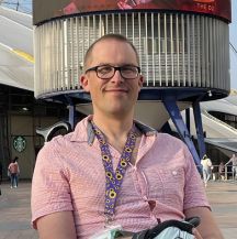 A head and shoulders shot of Sam who is smiling at the camera wearing glasses and a short sleeved shirt