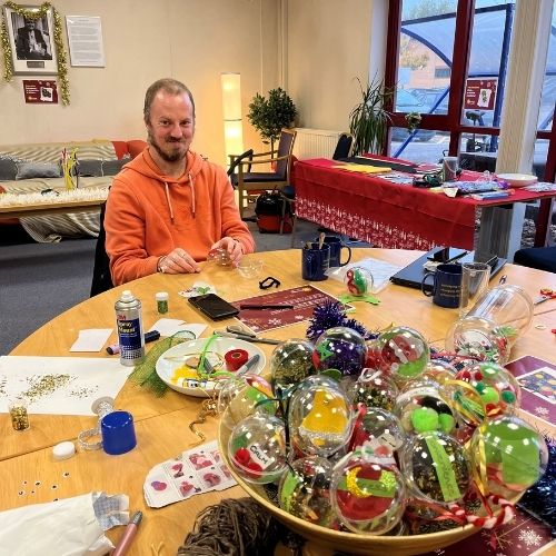 A crafter smiles at the camera sat at a craft table with crafts and baubles