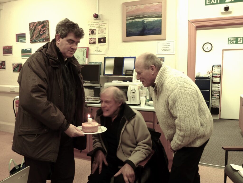 Brain sits between two colleagues blowing the candles out of a small cake