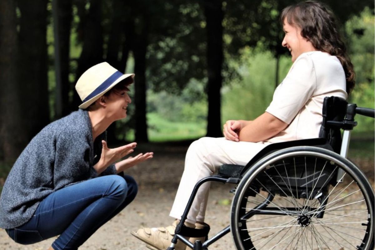 A person in hat crouches with hands outstretched in frint of a person in a wheelchair both smiling