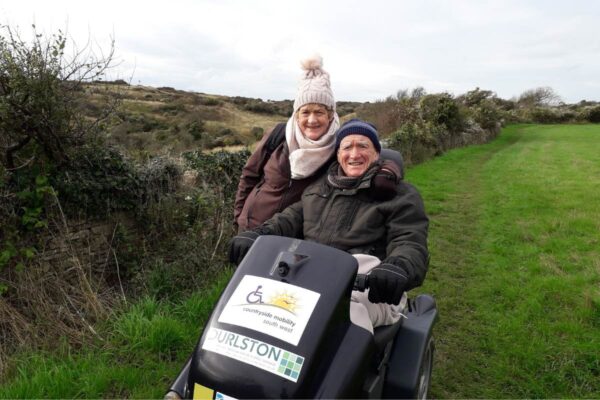 Denise Guest smiling alongside Barry Guest in a Countryside Mobility Tramper wrapped up warm on a grass pathway