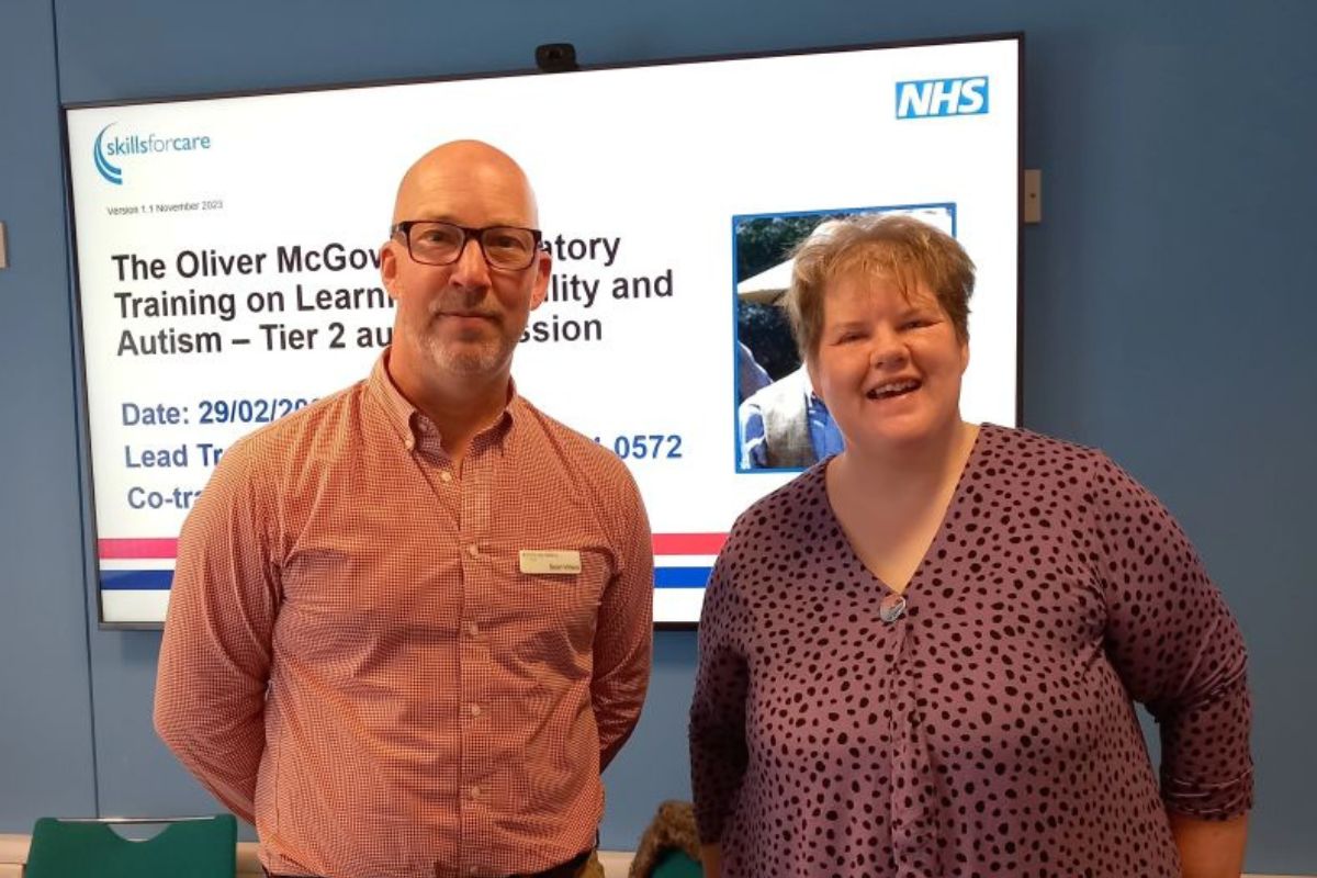 Heidi and Sean stand in front of a presentation screen smiling at the camera