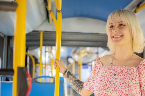 A smiling person stands on a bus holding to a hand rail