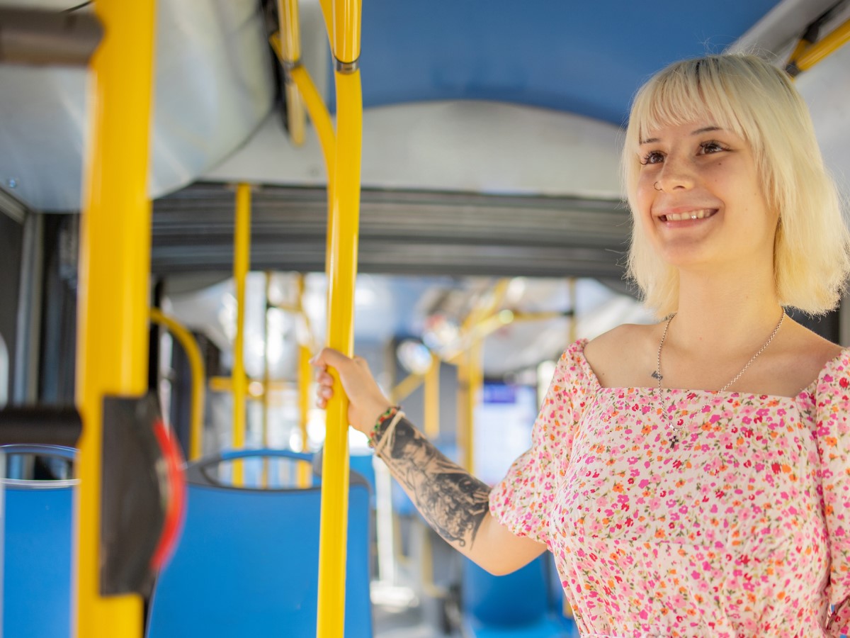 A smiling person stands on a bus holding to a hand rail