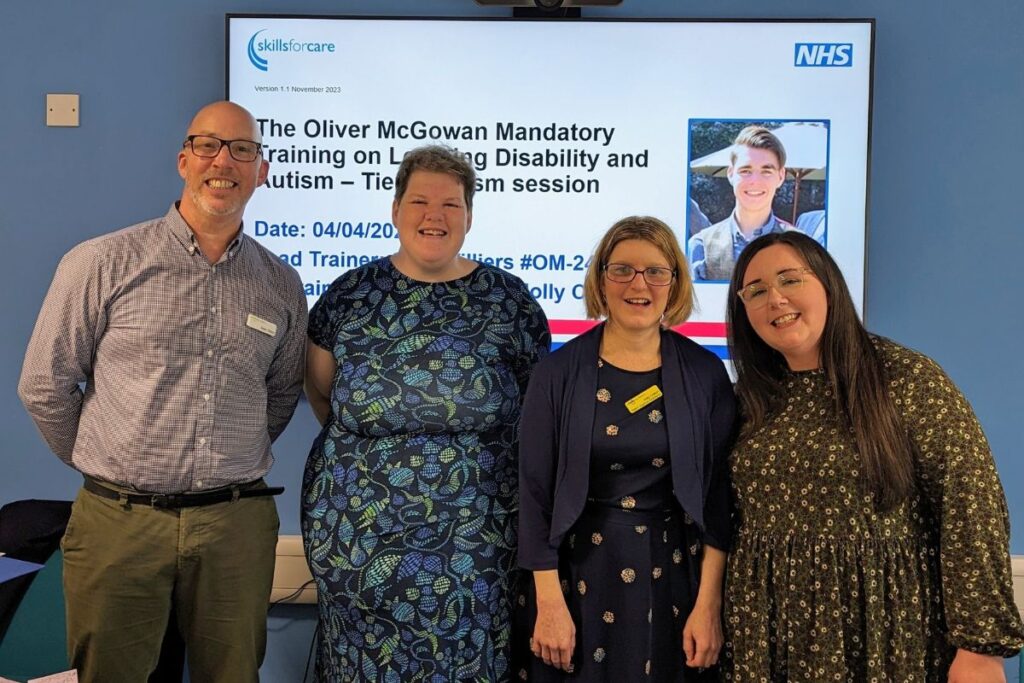 Sean, Heidi, Holly and Charlie stand smiling in front of an Oliver McGowan Training presentation slide