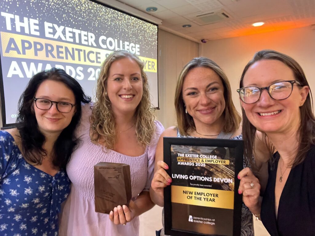 This image shows four women from LOD smiling and posing at the awards event. They are standing in front of a screen that reads: "The Exeter College Apprentice and Employer Awards 2025". Two of the women are holding awards. One holds a wooden trophy, and another holds a framed certificate reading: "Living Options Devon, New Employer of the year.