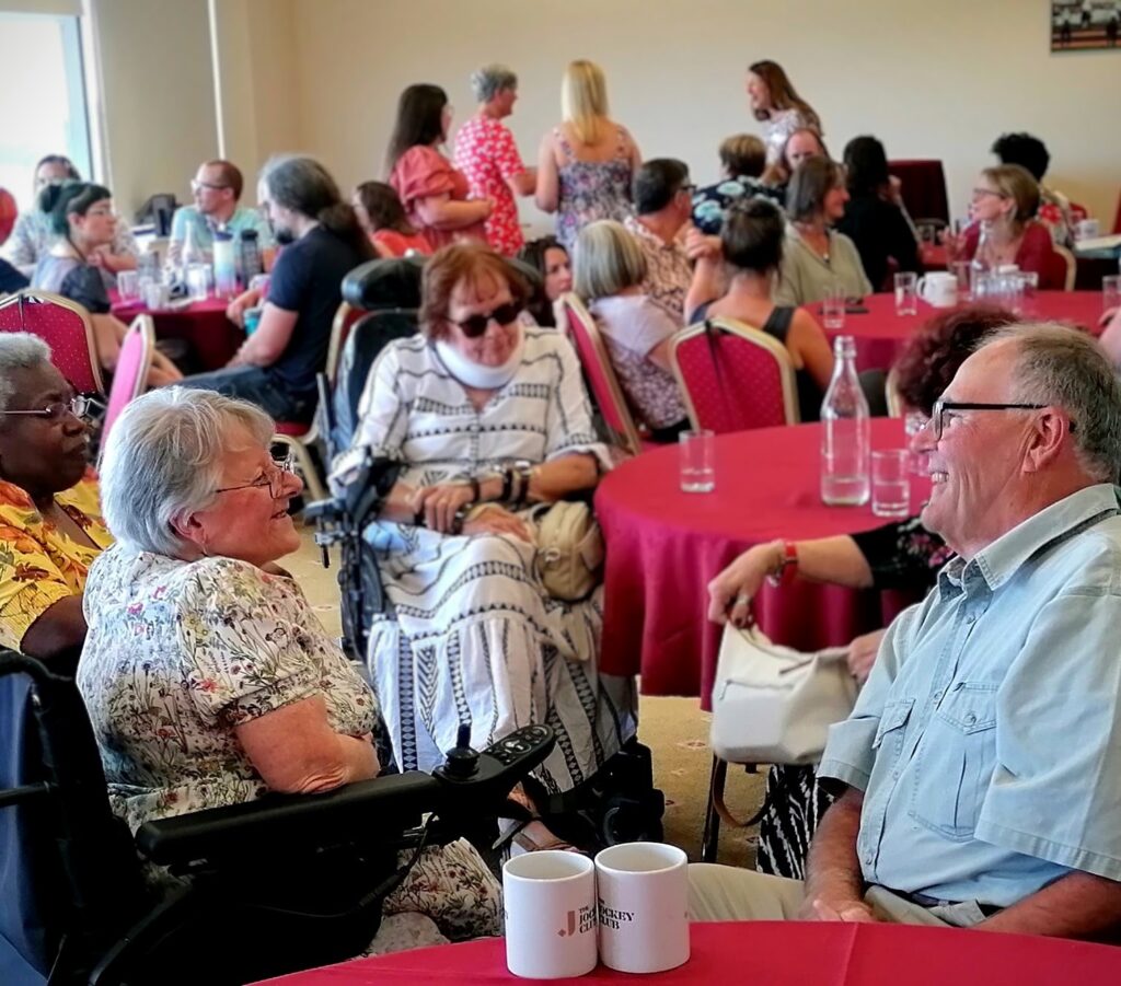 Overseeing the celebration. Lots of volunteers and staff members are sat around tables with various drinks. People are smiling and chatting.